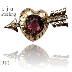 Könnte beinhalten: Eine antike Brosche aus Sterlingsilber in Herzform mit einem rosafarbenen Stein und einem goldenen Pfeil mit klaren Steinen. Die Brosche ist mit "Reja Sterling" und "1940" gekennzeichnet.