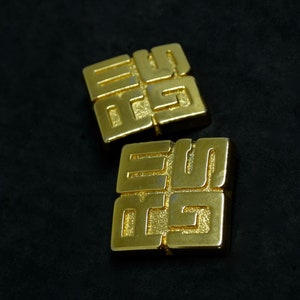 May include: Two gold-colored metal earrings with a geometric design. Each earring features a square shape with a raised pattern of interlocking squares.