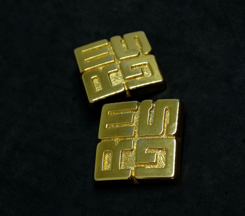 May include: Two gold-colored metal earrings with a geometric design. Each earring features a square shape with a raised pattern of interlocking squares.