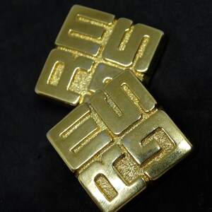 May include: A pair of gold-tone earrings with a geometric design. Each earring features a square shape with a raised pattern of interlocking squares.