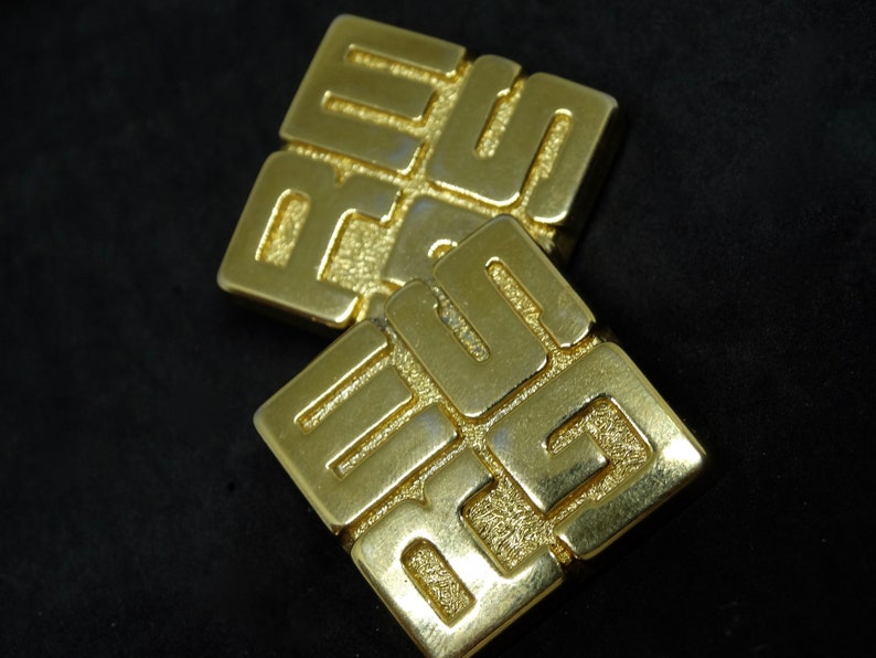 May include: A pair of gold-tone earrings with a geometric design. Each earring features a square shape with a raised pattern of interlocking squares.