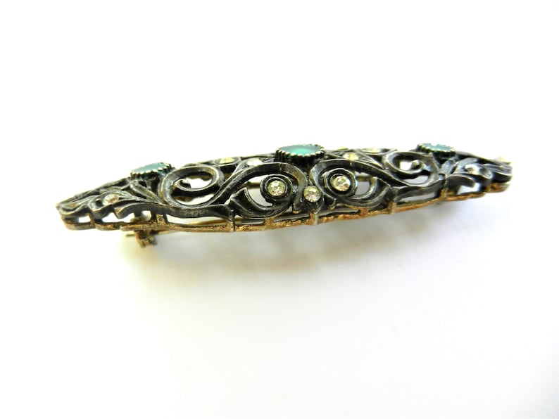 May include: A silver and gold filigree brooch with green and clear stones.