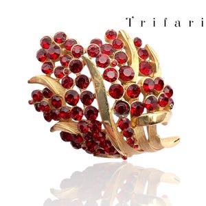 Op de afbeelding: Een vintage Trifari broche met rode strass-steentjes in een goudkleurig metalen ontwerp. De broche heeft een getextureerde, bladachtige vorm met een gebogen, gelaagd ontwerp. De merknaam Trifari is zichtbaar.
