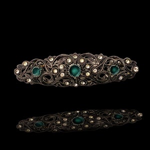 May include: A silver-toned hair clip with an ornate design featuring a floral pattern and green gemstones.
