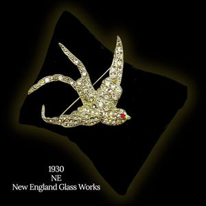 Pode incluir: Broche vintage de pássaro, adornado com strass transparentes e um olho vermelho. A peça é apresentada sobre um fundo preto. O texto "1930 NE New England Glass Works" é visível.