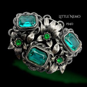Puede incluir: Broche plateado vintage con piedras preciosas verdes de talla esmeralda y redondas. El broche presenta un diseño floral con hojas y el texto "LITTLE NEMO 1940" sobre fondo negro. Joya única.