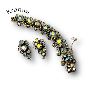 Può includere: Set di gioielli Kramer vintage con bracciale e orecchini abbinati. Il set presenta un design floreale con strass iridescenti e una montatura in metallo scuro. La parola "Kramer" è stampata in corsivo nero in alto a sinistra.