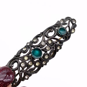 May include: A silver-toned hair clip with a delicate, ornate design featuring swirls and clear rhinestones. The clip has two green enamel stones.