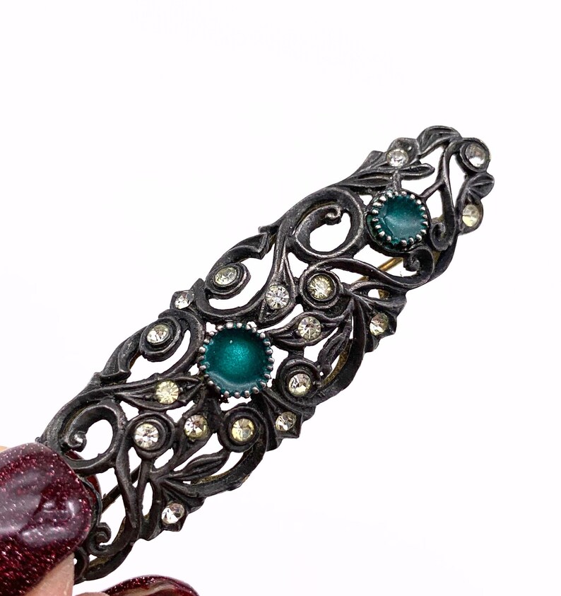 May include: A silver-toned hair clip with a delicate, ornate design featuring swirls and clear rhinestones. The clip has two green enamel stones.