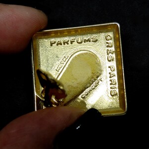 May include: Gold-colored metal perfume bottle cap with the words "Parfums Gres Paris" and "Metal Concept" engraved on it.