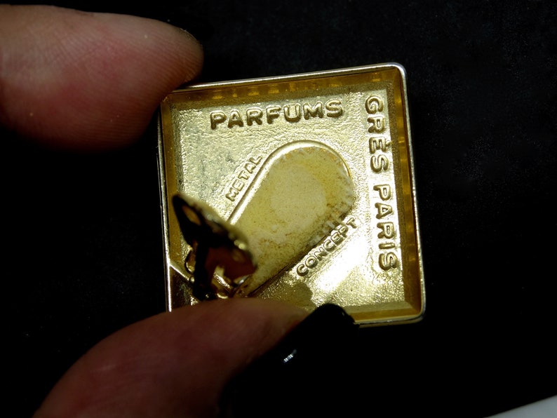 May include: Gold-colored metal perfume bottle cap with the words "Parfums Gres Paris" and "Metal Concept" engraved on it.