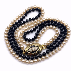 Può includere: Una collana a tre fili con perline color crema e nere alternate. La collana ha una chiusura dorata ornata di smalto nero e strass. Il gioiello è esposto su sfondo bianco. La collana è lunga circa 40 cm.