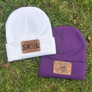 May include: A white knit beanie and a purple knit beanie, each featuring a rectangular brown faux leather patch secured with four gold-tone metal rivets. The white beanie patch displays the text SKOL, while the purple beanie patch shows a Viking.