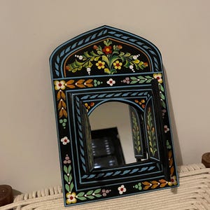 以下が含まれることがあります： 黒いアーチ型の鏡で、花の模様が施されています。 フレームは、黄色、オレンジ、白のカラフルな花と葉で飾られています。 鏡には2つのヒンジ付きシャッターがあり、ユニークな装飾要素を追加しています。 装飾的な家の装飾品です。