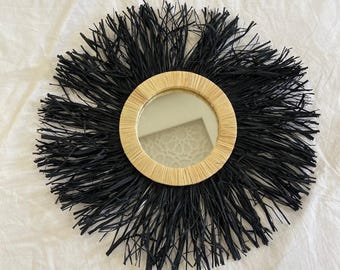May include: A round decorative mirror with a natural woven frame and a black fringe. The mirror's circular shape is accentuated by the contrasting textures of the smooth glass and the surrounding fringe.