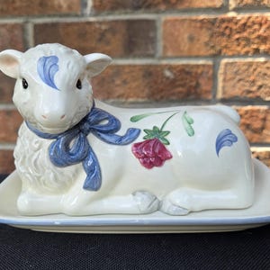 May include: A ceramic butter dish in the shape of a lamb, painted white with blue accents and a blue bow. The lamb has a floral design with a red flower and green leaves. The dish sits on a matching rectangular base.