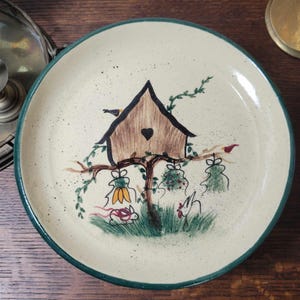 May include: A ceramic plate with a green rim features a hand-painted design of a birdhouse with a heart-shaped opening, surrounded by green vines and birds. Two bunnies and a rooster are also depicted on the plate.