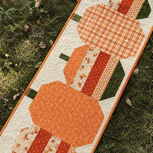 May include: A rectangular quilted table runner featuring a vertical arrangement of three pumpkins. The pumpkins are constructed from various orange, white, and green fabrics with plaid, polka dot, and leaf patterns, all set against a cream background.
