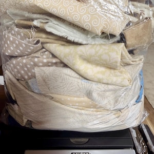 May include: A clear plastic bag filled with assorted beige and cream fabric scraps featuring various patterns including swirls, dots, and floral prints, resting on a digital scale that displays a weight of 2.38 pounds in clear text.