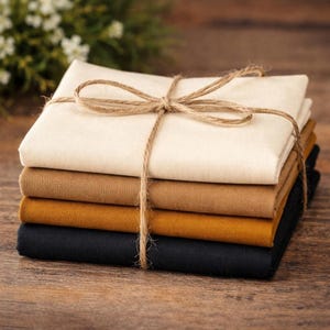 May include: A stack of four fabric squares in neutral colors: cream, tan, mustard, and black. The squares are neatly folded and tied together with twine, forming a neat bundle. The fabric appears to be cotton or linen, suitable for quilting or crafting.