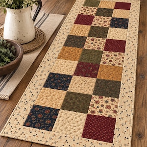May include: A rectangular quilted table runner featuring a patchwork design of alternating squares in shades of burgundy, navy blue, olive green, and cream. The runner is bordered by a cream fabric with a small repeating floral and leaf pattern.
