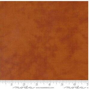 May include: A close-up of a piece of fabric with a mottled, textured appearance. The fabric is primarily a warm, earthy brown color with darker areas creating a subtle pattern. A ruler is at the bottom of the image, with the word "moda" printed on it.