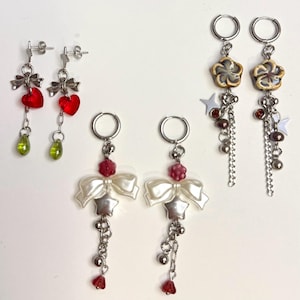 May include: A collection of dangle earrings with various designs. One pair features red heart charms and green teardrop beads. Other pairs include white bows, star accents, and flower-shaped charms. All earrings have silver-tone metal details.