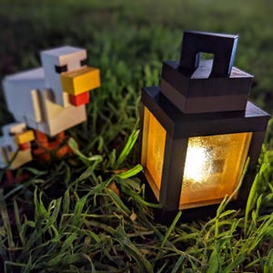 May include: A black lantern with a lit candle inside, sitting on green grass. Two white Minecraft chicken figurines are in the foreground.