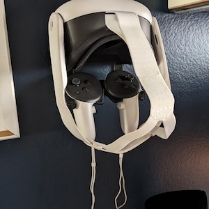 May include: A white virtual reality headset with a white strap and two black controllers hanging on a wall.