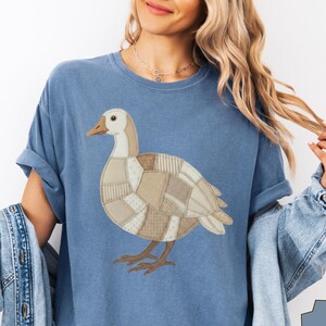 Peut inclure: Un t-shirt bleu jean avec un motif central représentant un canard composé de divers motifs en patchwork beige et brun clair. Le canard a une tête claire, un bec brun et des pattes brunes. Le t-shirt a des manches courtes et un col rond.