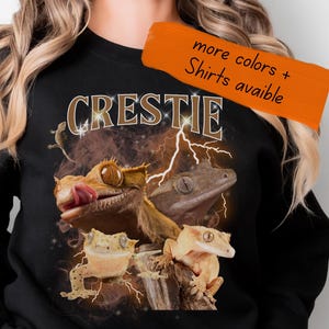 Peut inclure: Sweat-shirt noir avec un motif de geckos à crête et le mot "CRESTIE". Le design utilise des tons marron et jaune avec des éclairs. Une bannière orange en haut à droite indique "more colors + Shirts avaible".