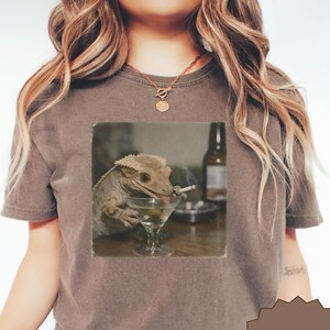 Può includere: Una t-shirt marrone a maniche corte con una stampa grafica di un geco crestato che tiene una sigaretta accanto a un bicchiere da martini. La maglietta presenta un girocollo e una vestibilità rilassata con un'immagine in stile fotografico.