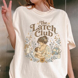 May include: An ivory t-shirt featuring a graphic print with the text "The Latch Club" above an illustration of a person breastfeeding an infant, flanked by two breast pumps, all surrounded by a decorative floral wreath in muted earth tones.