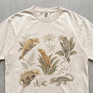 Puede incluir: Una camiseta de manga corta color marfil con un estampado gráfico detallado de cuatro geckos crestados en varias posturas entre ilustraciones botánicas de helechos, hojas y pequeñas flores situadas en el centro del pecho.