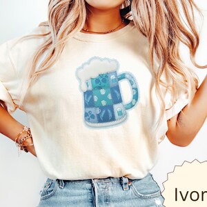 Op de afbeelding: Een ivoorkleurig T-shirt met korte mouwen en een centrale afbeelding van een bierpul. De pul is ontworpen met een blauw patchworkpatroon met diverse kleine iconen zoals korenaren, bekroond met een dikke laag wit schuim.