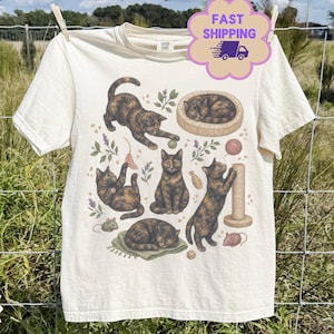 Pode incluir: Uma t-shirt de manga curta na cor marfim com uma estampa gráfica de vários gatos tartaruga em várias poses, incluindo a dormir numa cama, a brincar com brinquedos e a usar um arranhador, rodeados por pequenas ilustrações botânicas.