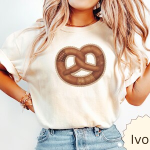 May include: An ivory short-sleeved t-shirt featuring a large, centered graphic of a brown pretzel. The pretzel design has a scalloped edge and incorporates subtle patterns within its shape. The shirt is paired with blue denim jeans.