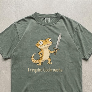 Peut inclure: Un t-shirt à manches courtes vert mousse avec un motif représentant un lézard jaune tenant une épée argentée. Sous le lézard, le texte I require Cockroaches est inscrit dans une police sombre. Le t-shirt a un col rond classique.