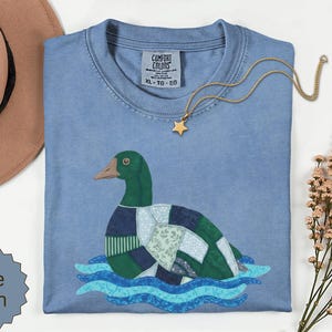 Pode incluir: Uma t-shirt azul ganga com um gráfico de um pato-real desenhado com um padrão de retalhos de vários tecidos verdes e azuis, a flutuar sobre ondas de água azuis estilizadas. A t-shirt tem uma etiqueta Comfort Colors na gola.