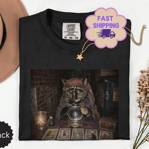 Pode incluir: Uma t-shirt preta com uma estampa gráfica de um gato vestido de cartomante com uma bola de cristal e cartas de tarot. A t-shirt tem uma etiqueta Comfort Colors no colarinho e é exibida ao lado de um chapéu bege e flores secas.