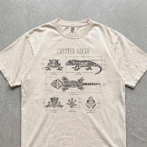 Puede incluir: Una camiseta de manga corta color marfil con un estampado gráfico negro de un gecko crestado con varias vistas anatómicas, incluyendo perspectivas frontal, lateral, superior y trasera, etiquetadas con medidas técnicas en milímetros y el texto Crested Gecko.