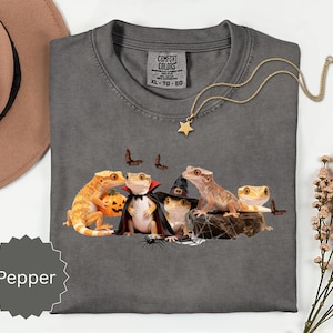 Puede incluir: Una camiseta gris carbón con un gráfico de cuatro geckos crestados disfrazados de Halloween, incluyendo un vampiro y una bruja, con una calabaza, murciélagos y una telaraña. La camiseta tiene una etiqueta Comfort Colors de talla XL-TG-EG.