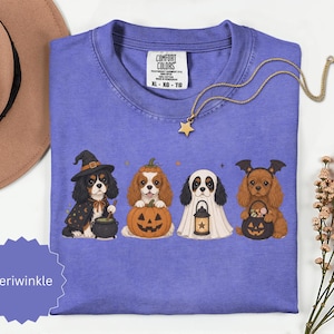 May include: A periwinkle t-shirt featuring a graphic of four Cavalier King Charles Spaniels dressed for Halloween. The dogs are depicted as a witch with a cauldron, a pumpkin, a ghost with a lantern, and a bat with a candy bucket.
