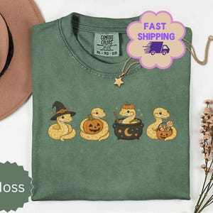 May include: A moss green short-sleeve t-shirt featuring a graphic of four snakes in various Halloween-themed poses, including one wearing a witch hat, one with a jack-o'-lantern, one in a cauldron, and one holding a candy bucket with a pumpkin.