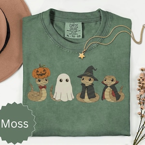 May include: A moss green t-shirt with a Comfort Colors label in size XL. The graphic features four snakes: one wearing a pumpkin, one as a ghost, one in a witch hat and cloak, and one in a vampire cape. A gold star necklace rests on the neckline.