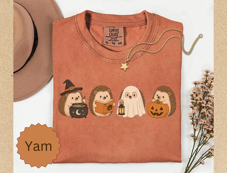 May include: A rust-colored t-shirt featuring a graphic of four hedgehogs in Halloween costumes: one with a witch hat and cauldron, one reading a book, one dressed as a ghost holding a lantern, and one holding a jack-o'-lantern pumpkin.