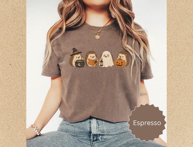 May include: An espresso-colored short-sleeve t-shirt featuring a graphic print of four hedgehogs in Halloween costumes. One hedgehog wears a witch hat with a cauldron, one reads a book, one is dressed as a ghost with a lantern, and one holds a pumpkin.