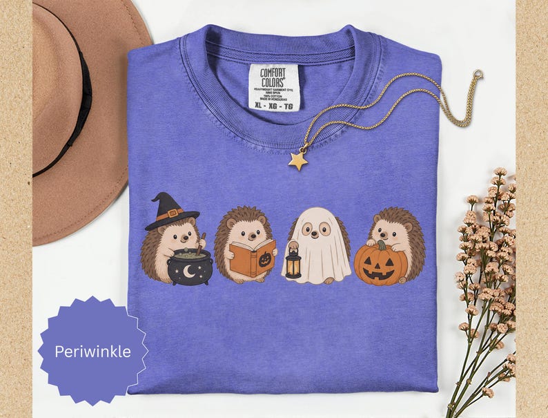 May include: A periwinkle t-shirt featuring a graphic of four hedgehogs dressed for Halloween: one with a witch hat and cauldron, one reading a book, one wearing a ghost sheet with a lantern, and one holding a jack-o'-lantern pumpkin.