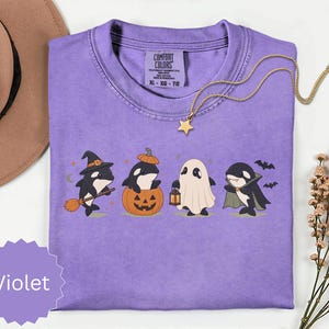 May include: A violet short-sleeved t-shirt featuring a graphic of four orcas dressed in Halloween costumes: a witch with a broom, an orca in a pumpkin, a ghost holding a lantern, and a vampire with a cape. The shirt has a crew neck and a Comfort Colors label.