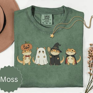 Peut inclure: Un t-shirt vert mousse avec un motif représentant quatre dragons barbus en costumes d'Halloween : un avec une citrouille, un en fantôme, un en sorcière avec un balai et un avec une cape. L'étiquette Comfort Colors indique la taille XL.